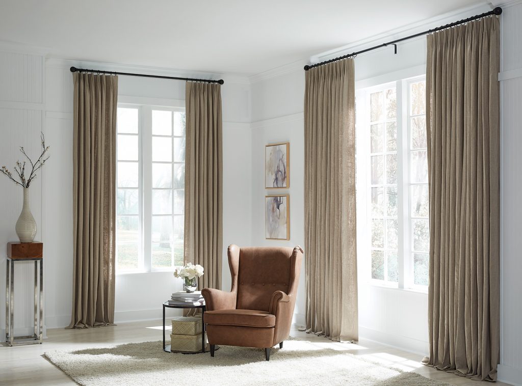 A simple room in whites and beiges with an armchair and small table nestled in a corner between two tall windows.