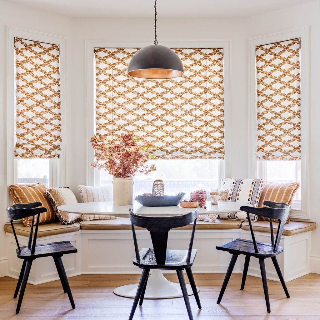 A kitchen nook with bench seating in a bay window featuring Flat Roman Soft Shades in a large scale curved line pattern in browns on an off-white background.