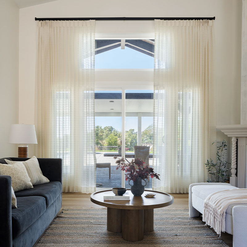 A high-ceiling living room with custom white sheer drpaery over tall glass doors.