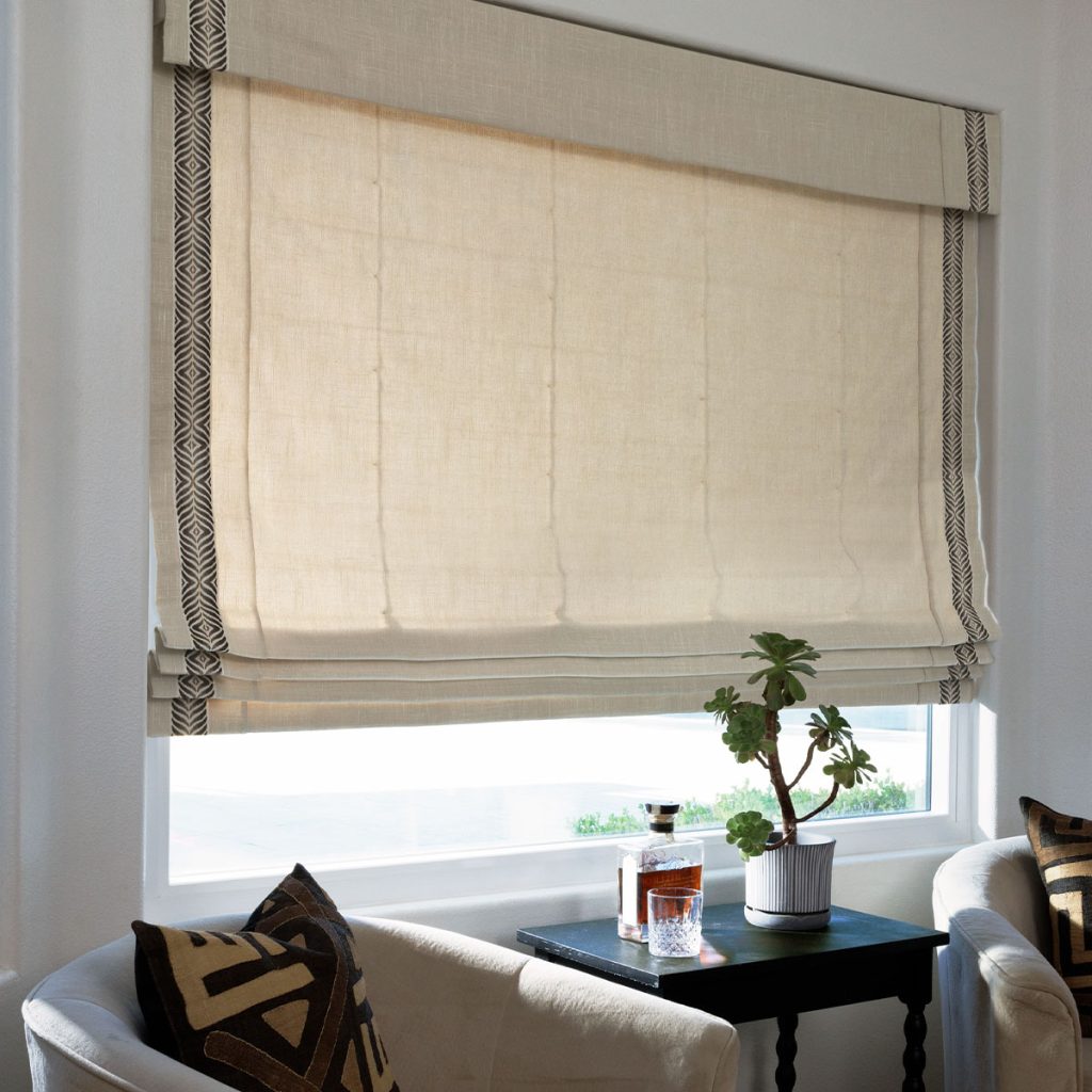 A beige Flat Roman Shade positioned off the back of the headrail with vertical trim in a sitting area.